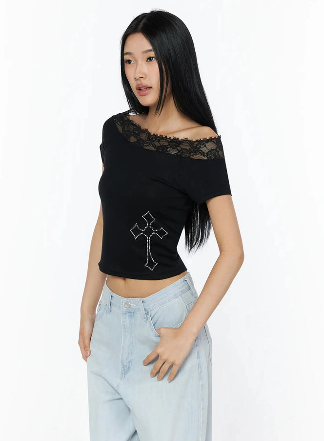 Rhinestone Cross Lace Trim Off-Shoulder Top IL516 Black three-quarter front shot model standing wearing fitted crop top with lace neckline and rhinestone cross motif styled with light blue jeans