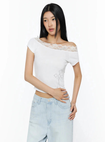Rhinestone Cross Lace Trim Off-Shoulder Top IL516 White three-quarter front shot model standing hand on waist lace off-shoulder detail and rhinestone cross visible styled with light blue wide-leg jeans