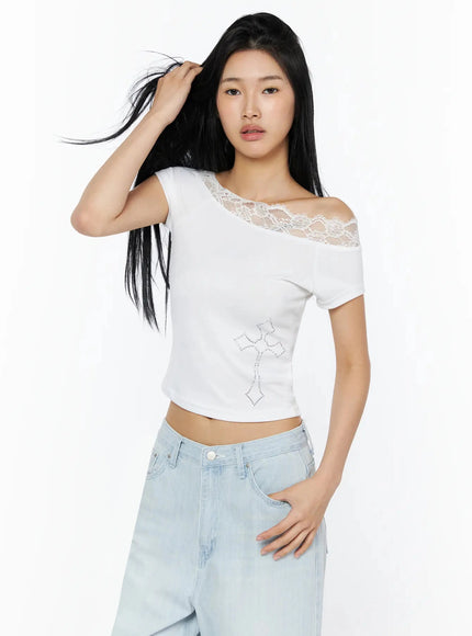 Rhinestone Cross Lace Trim Off-Shoulder Top IL516 White three-quarter front shot model hand touching hair showcasing lace neckline rhinestone cross motif and cropped silhouette with light blue wide-leg jeans