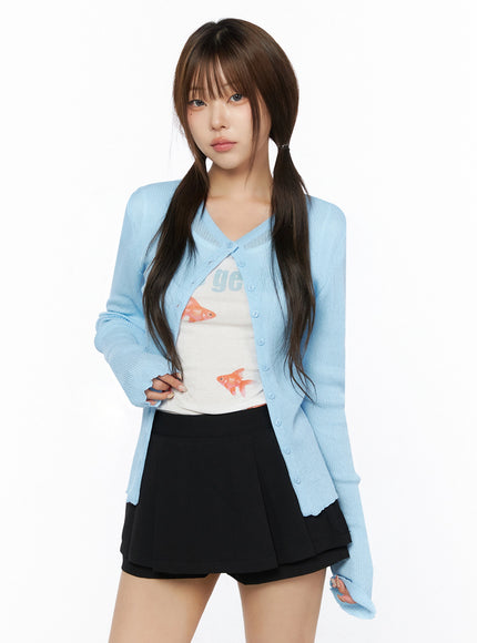 Ribbed Round Neck Cardigan IL515 Light blue three-quarter front model shot standing upright, ribbed knit cardigan over goldfish tank and black pleated mini skirt