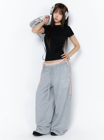 Ribbed Short Sleeve Tee IL515 Black full-body lifestyle shot model wearing fitted ribbed crop tee with silver headphones and bag styled with gray wide-leg sweatpants
