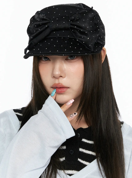 Ribbon Cubic Hat CL517 Black close-up front view on model hand touching chin, rhinestone-studded satin cap with large bow, styled with striped knit vest