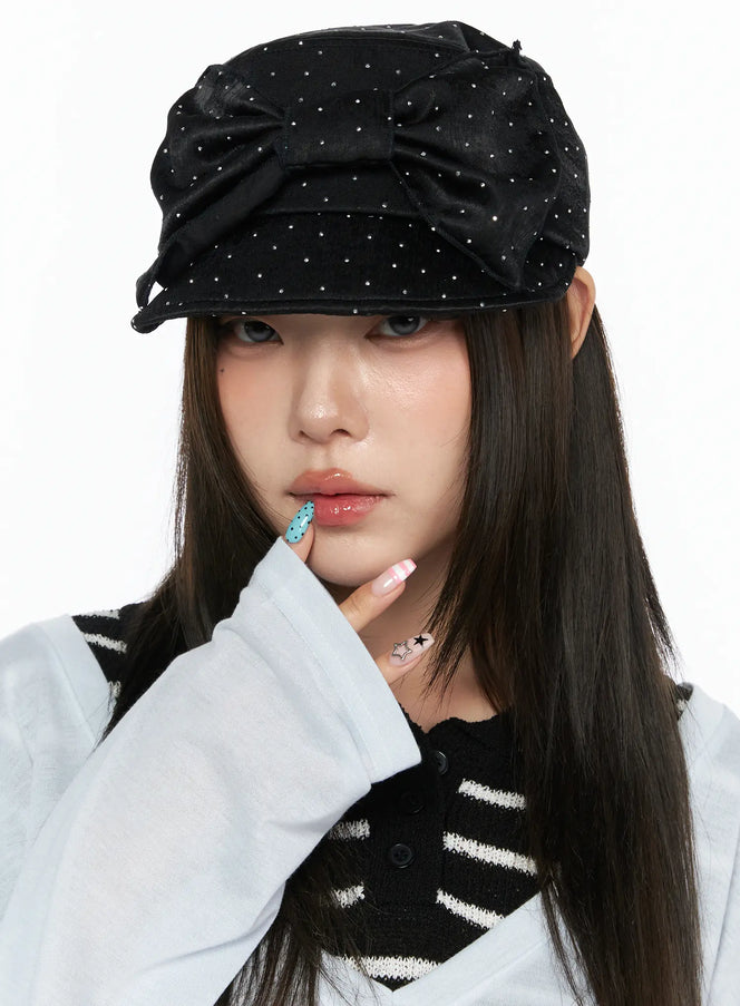 Ribbon Cubic Hat CL517 Black close-up front view on model hand touching chin, rhinestone-studded satin cap with large bow, styled with striped knit vest