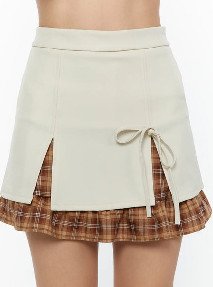 Ribbon Pleated Mini Skirt CU526 Light beige front close-up detail shot showing ribbon bow tie center split and layered brown plaid pleated underlayer