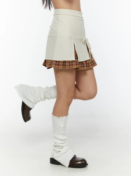 Ribbon Pleated Mini Skirt CU526 Light beige side shot on model with leg raised showing split hem and brown plaid pleated underlayer with white leg warmers