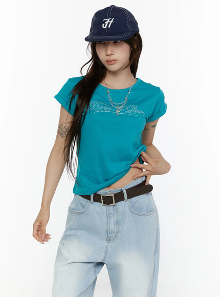 Rolled-Up Graphic Slim Fit Tee CL507 Blue front shot on model with cap and layered necklace hand pulling hem rhinestone script graphic and slim cotton fit visible