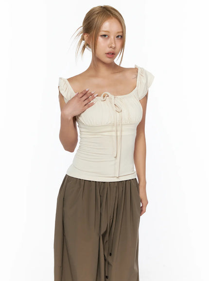 Ruched Off-Shoulder Bow Detail Top CU524 light beige three-quarter front shot on model hand on chest highlighting ruffle sleeves square neckline and bow tie