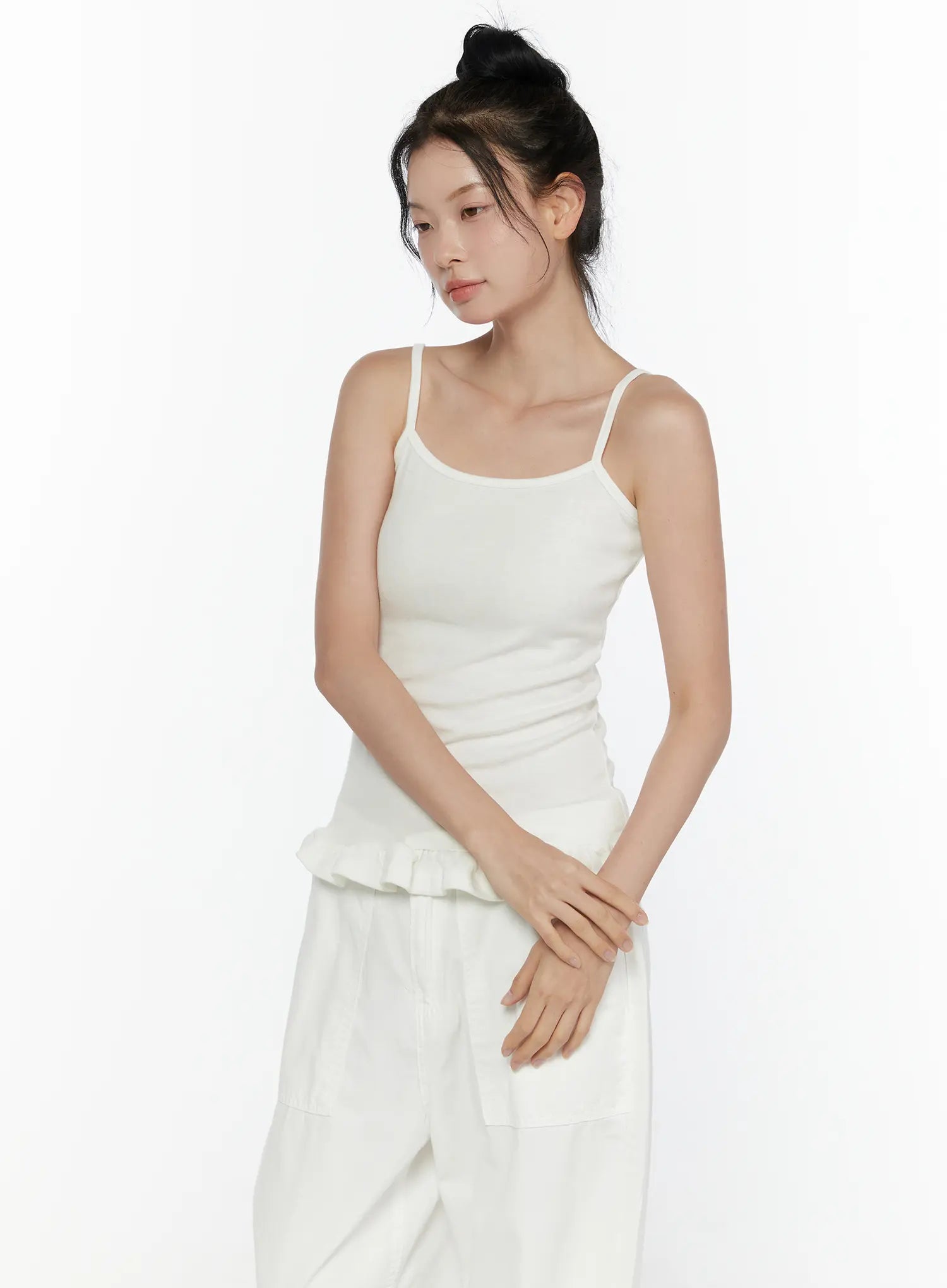 Ruffle Detail Cami Top CL521 White three-quarter front shot on model, spaghetti strap cami with ribbed knit texture, ruffled peplum hem, hands clasped at waist, paired with white wide-leg pants