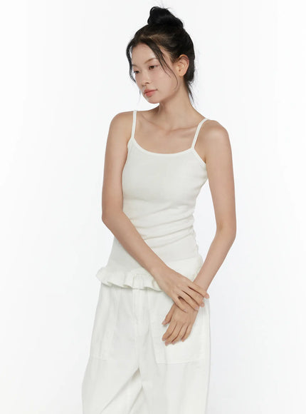 Ruffle Detail Cami Top CL521 White three-quarter front shot on model, spaghetti strap cami with ribbed knit texture, ruffled peplum hem, hands clasped at waist, paired with white wide-leg pants