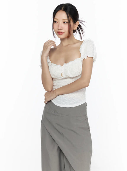 Ruffle Detailed Blouse CL527 White three-quarter front model shot with hand raised near chin, showcasing puff sleeves and scalloped neckline, styled with gray trousers