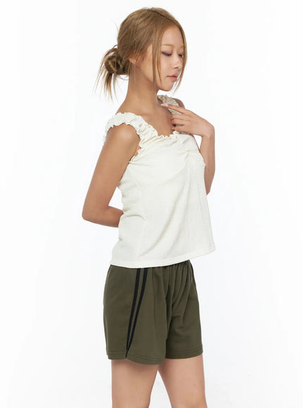 Shirred Ruffle Tank Top CU524 Light beige side close-up on model touching ruffle shoulder strap with hand paired with olive side-stripe shorts