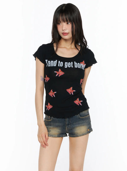 Slim Fit Graphic Tee CU530 Black front shot on model with blunt bangs, hands relaxed at sides, paired with distressed denim shorts, showing fitted silhouette and goldfish print