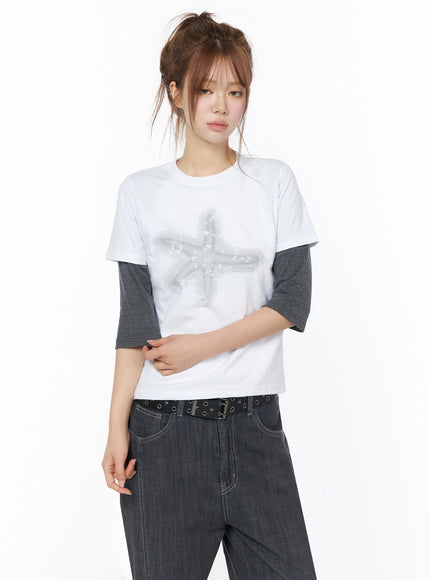 Star Graphic Tee IU525 White three-quarter front model shot with star graphic layered over gray long-sleeve tee hands clasped at waist styled with belted dark jeans