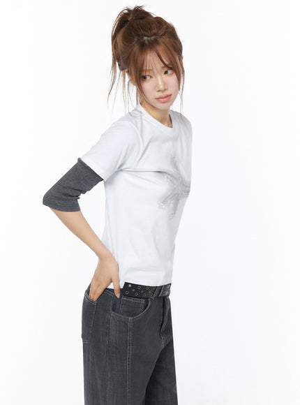 Star Graphic Tee IU525 White side model shot showing fitted silhouette layered over gray long-sleeve tee hand on hip styled with dark wide-leg jeans and eyelet belt