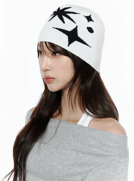 Star Knit Beanie IL506 White three-quarter on-model close-up, female model slight downward gaze, black starburst intarsia pattern on white knit beanie with grey off-shoulder top