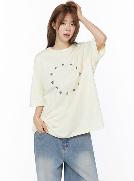 Star Ring Graphic Oversized Tee IL502 Light beige front shot model hand raised touching hair relaxed pose showing oversized boxy cut with star graphic and jeans