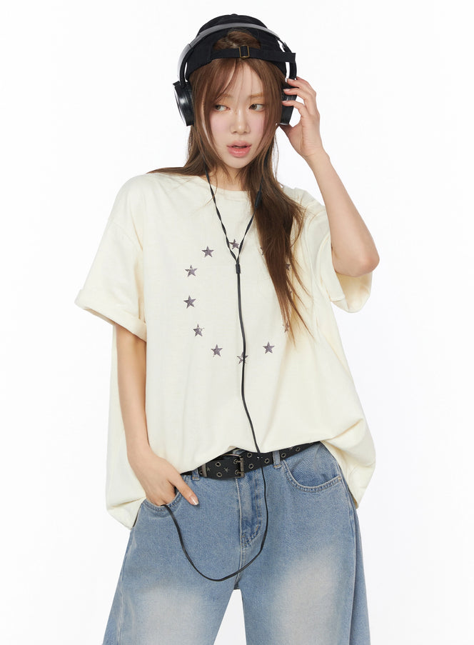 LEWKIN Star Ring Graphic Oversized Tee IL502 Light beige front crop shot model wearing headphones hand raised oversized cotton tee with scattered star print
