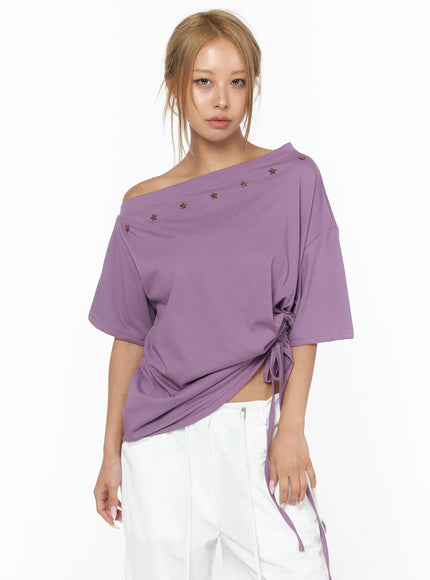Star Studded Asymmetric Drawstring Tee CU524 Purple front shot on model with direct gaze, off-shoulder asymmetric collar with bronze star studs, side drawstring ruching, white wide-leg trousers