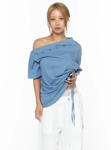 Star Studded Asymmetric Drawstring Tee CU524 Blue front shot on model with one-shoulder off, bronze star studs at neckline, ruched gathered hem with drawstring tie, white wide-leg pants