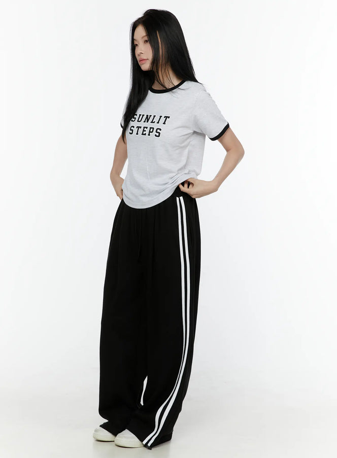Stripe Detail Sweatpants IL506 Black side-angle full-body model shot, styled with grey graphic tee, wide-leg fit with white double side stripes, white sneakers