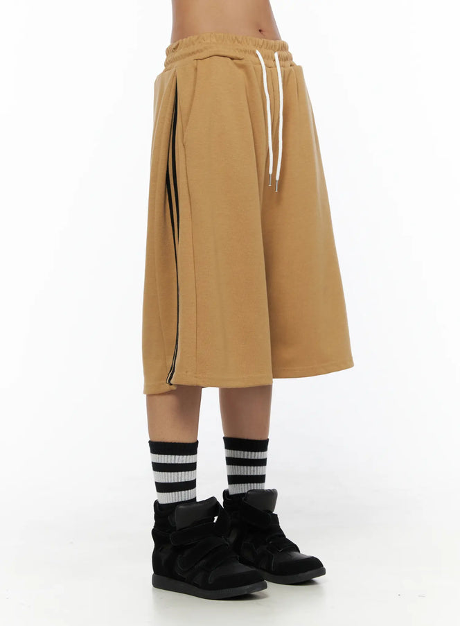 Striped Bermuda Shorts CL503 Beige side view on model showing white drawstring waistband, black side stripe detail and wide-leg silhouette with black sneakers