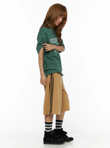 Striped Bermuda Shorts CL503 Beige side full-body on model looking down, green oversized tee tucked slightly, black side stripes visible, striped socks and black sneakers