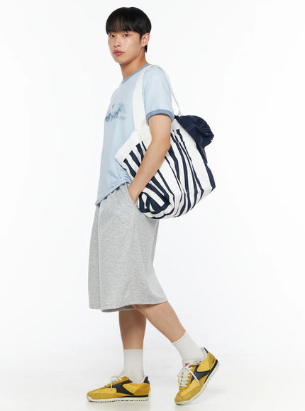 Striped Canvas Tote Bag IL514 Dark blue three-quarter shot on male model holding bag at side, navy stripe large canvas tote, white graphic tee and light grey shorts