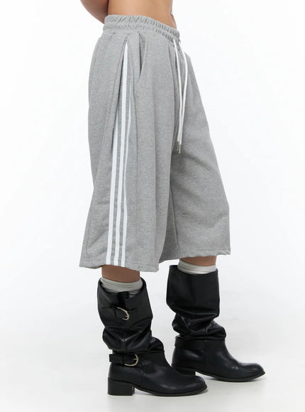 Track Bermuda Shorts CU523 Gray side close-up on model highlighting three white side stripes, pleated waist, and wide-leg silhouette with black boots
