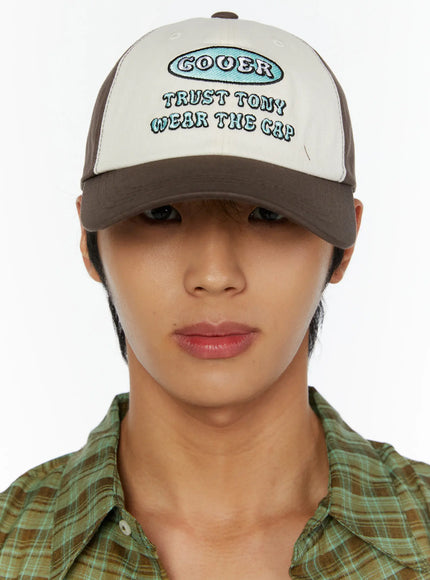 Two-Tone Graphic Cap IL515 Dark brown close-up front model shot, male model facing camera showing teal COOER embroidered oval logo and text on cream panel