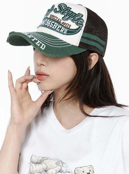 Vintage Graphic Cap IL506 Green side-angle model shot, woman gazing down with hand near chin, distressed trucker cap with brown mesh panel