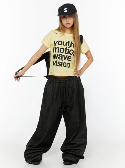 Youth Graphic Short Sleeve Tee IL524 Yellow full-body front view on model wearing black cap and holding backpack strap, styled with wide-leg dark denim