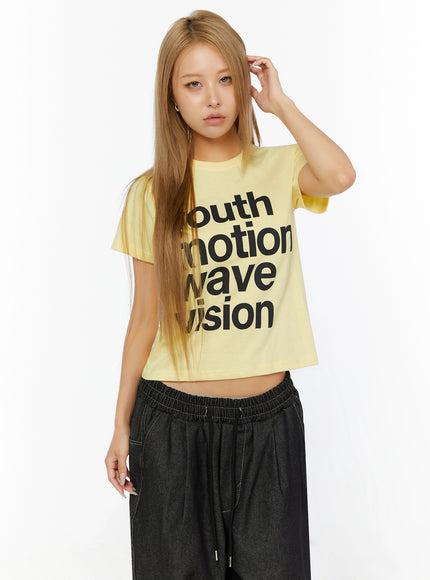 Youth Graphic Short Sleeve Tee IL524 Yellow front view on model with hand touching hair, cropped fit with black graphic print styled with dark drawstring pants