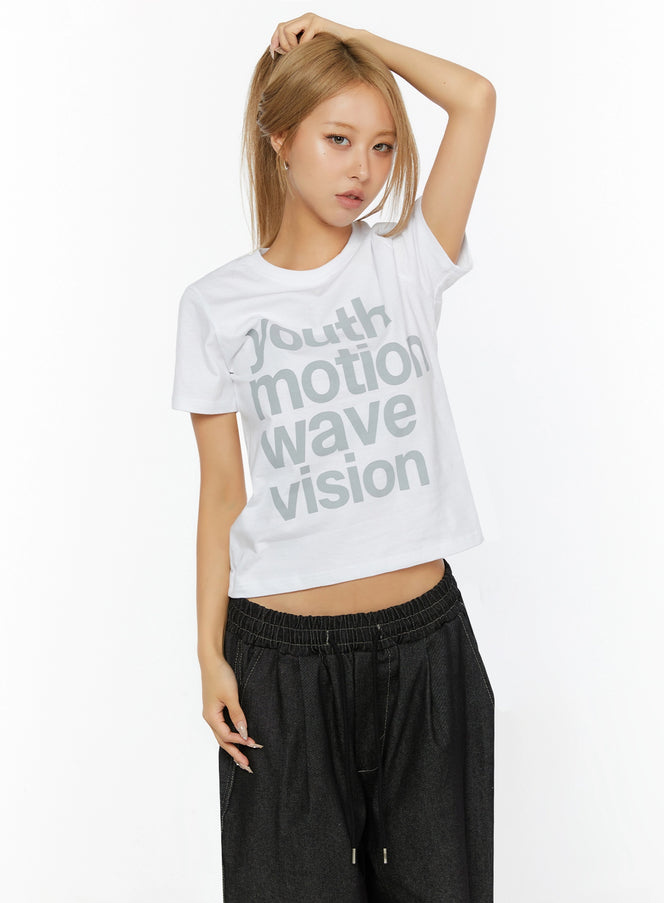 Youth Graphic Short Sleeve Tee IL524 White front view on model with hand raised to head, styled with wide-leg dark drawstring pants, cropped cotton tee