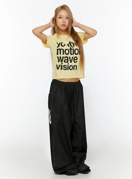 Youth Graphic Short Sleeve Tee IL524 Yellow front view on model with both hands raised behind head, cropped silhouette with wide-leg dark denim pants