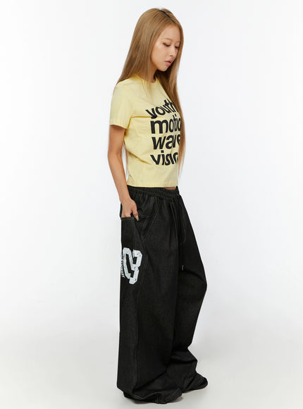 Youth Graphic Short Sleeve Tee IL524 Yellow side view on model looking down, showing cropped silhouette and sleeve detail, styled with dark wide-leg denim