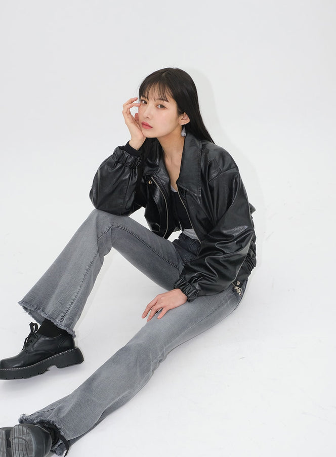 LEWKIN Faux Leather Crop Jacket J19 in black, model seated on floor, three-quarter view, glossy faux leather with banded hem and cuffs, styled with grey flare jeans and chunky boots.