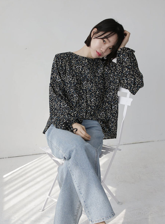 Shirring Blouse in black floral print, full-body seated shot on model in chair, relaxed wide fit with long sleeves, shirring detail across front, styled with light blue wide-leg jeans.