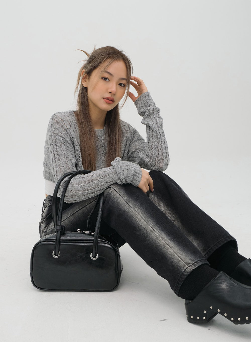 LEWKIN Black Jeans C1002 in washed black, model seated on floor with knees up showing faded leg panels, wide-fit cotton denim, paired with grey cable-knit crop and black mini bag.