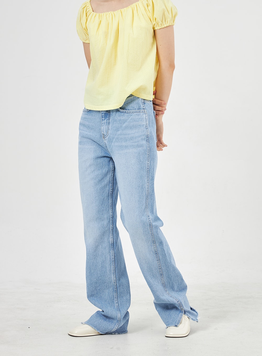 LEWKIN Light Wash Jeans OA310 in light blue, angled front-lower shot on model, wide-fit washed cotton denim with side slit hem, paired with yellow puff-sleeve top and white loafers.