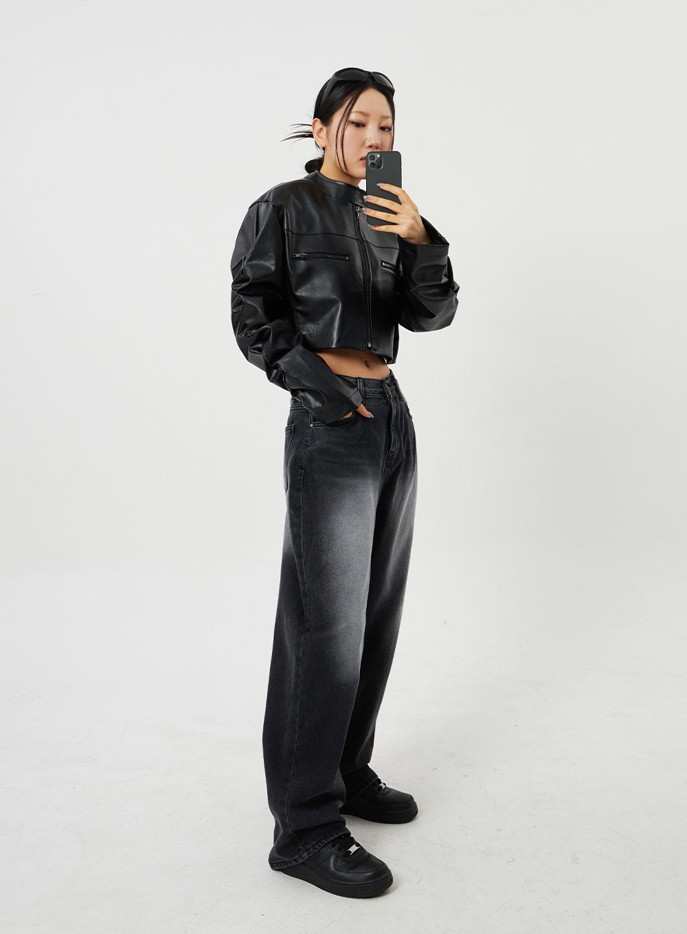 Mid-Rise Baggy Jeans CF328 in black, full-body side view on model taking mirror selfie, wide-fit faded denim styled with cropped faux-leather biker jacket and chunky sneakers.