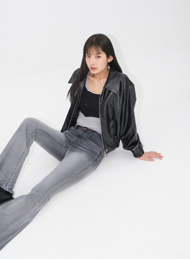 Faux Leather Crop Jacket J19 in black, model seated on floor, front-facing view, collar open revealing layered black crop top and grey tube top, paired with grey flare jeans.