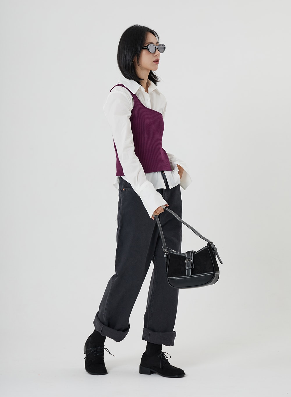 Maxi Denim Pants ON23 in charcoal grey, side-angle full-body shot on model holding black shoulder bag, wide-fit denim with cuffed hem, paired with white shirt, purple vest, and black oxfords.
