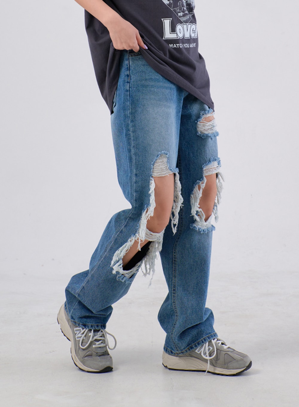 Ripped Knee Jeans IA305 in medium blue, side view on model mid-stride, showing wide-leg silhouette and deep frayed knee rips with dangling threads, worn with grey sneakers.