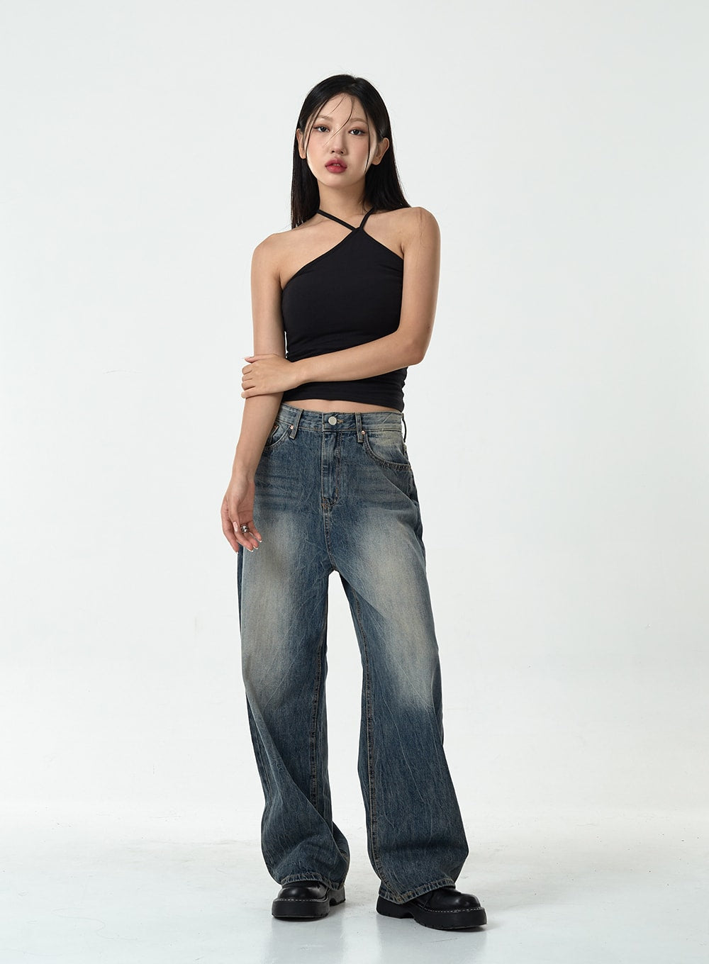 Washed Denim Pants CG10 in dark blue, full front view on model with arms crossed, wide-leg washed cotton denim with faded finish, paired with black cross-neck halter top and black loafers.