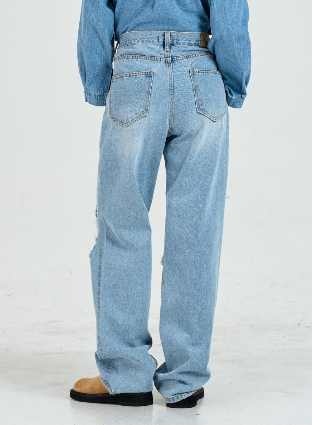 Ripped Maxi Denim Pants Unisex CO14 in light blue, back view on model, wide-fit cotton denim showing dual back pockets and visible rip on left knee, paired with denim shirt.