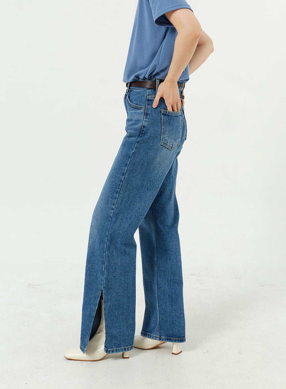 Side Slit Jeans OM308 in mid blue, side view close-up on model, wide-leg cotton denim highlighting the front side slit detail at hem, brown belt, white boots.