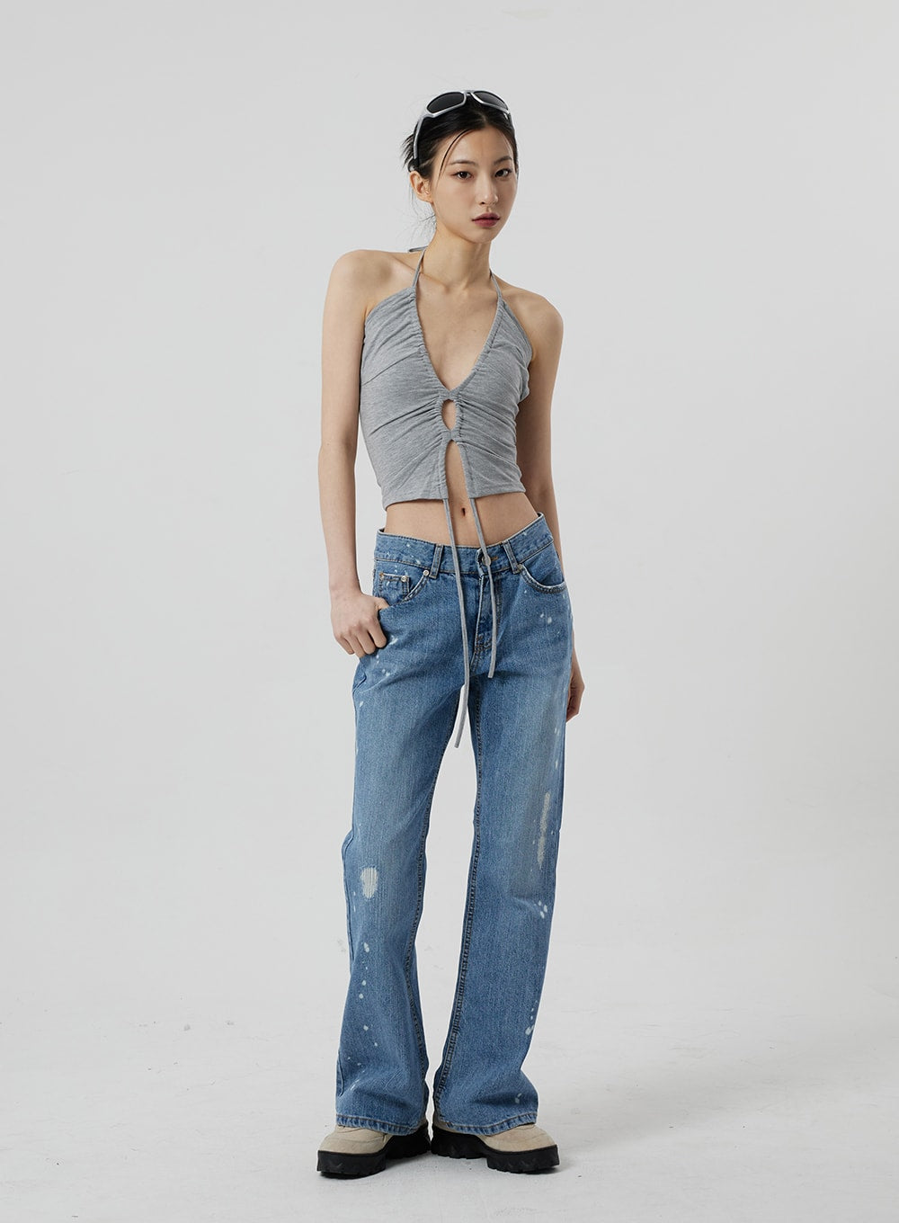 Low Rise Bootcut Jeans CF309 in blue denim, full-length front view on model, hand tucked in pocket, low waist with paint-splatter cotton denim, bootcut silhouette, paired with sleeveless grey halter crop top.