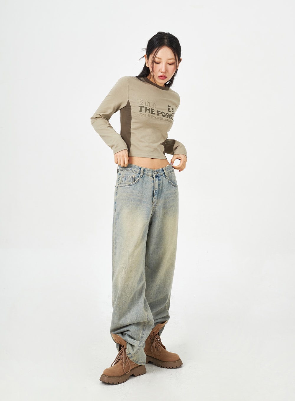 Light Blue Baggy Jeans CM323 in light blue, full-body front shot on model adjusting waistband, low-rise wide-fit denim with heavy washed fading, paired with beige graphic crop top.