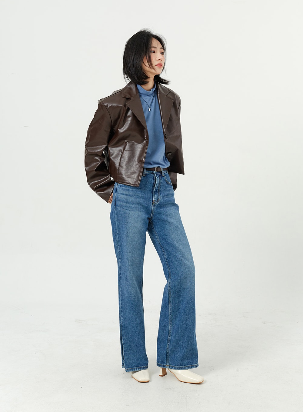 Side Slit Jeans OM308 in mid blue, full-length three-quarter angled shot on model, wide-fit cotton denim with side slit hem, styled with brown leather jacket, white boots.