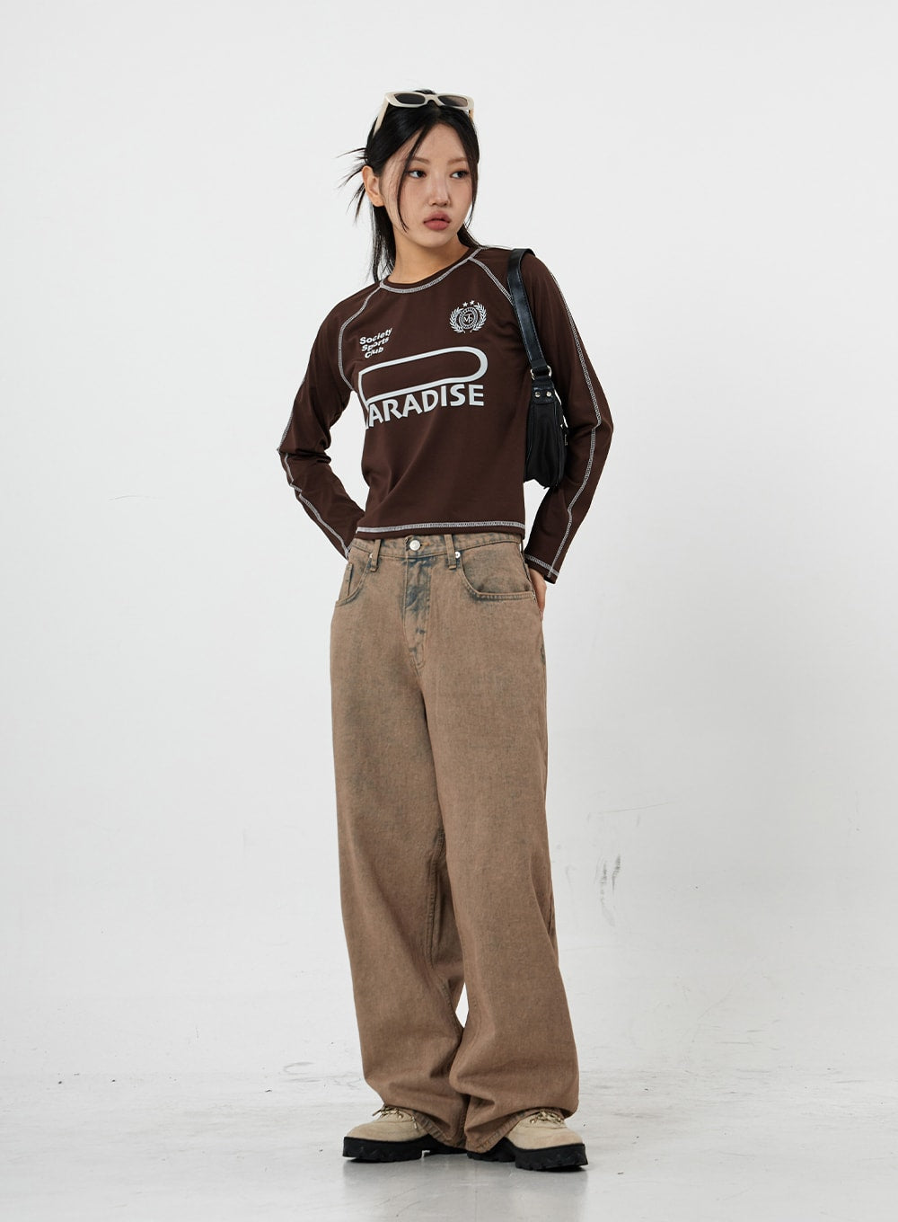 Raemon Wide Jeans Unisex CS21 in brown acid-wash, full-length front view on model with arms back, relaxed wide-leg fit, paired with brown graphic long-sleeve and crossbody bag.